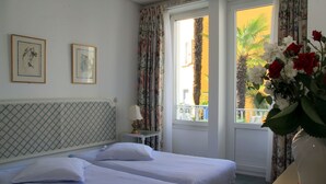 A hotel room with two beds, a window with curtains, a vase with flowers, and framed pictures on the wall. - Piccolo Hotel (Muralto)