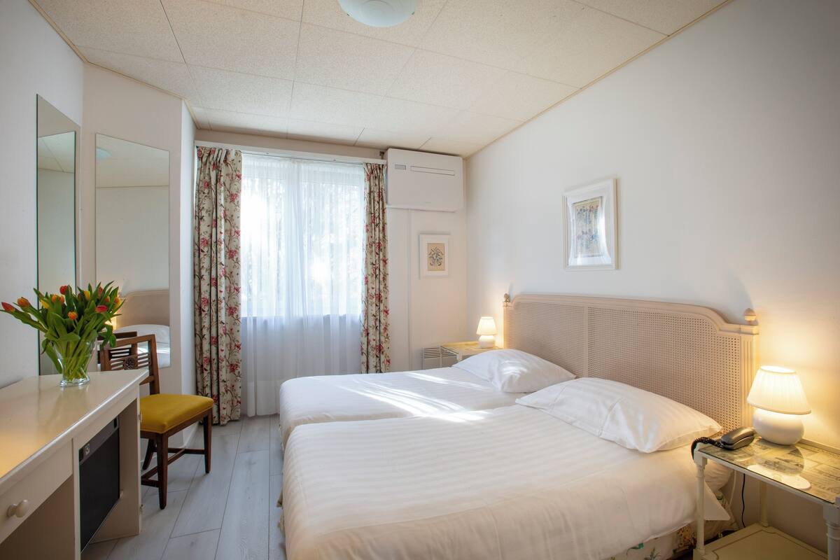 A hotel room with two beds, a desk with a chair, a mirror, a window with curtains, and a bedside table with a lamp.