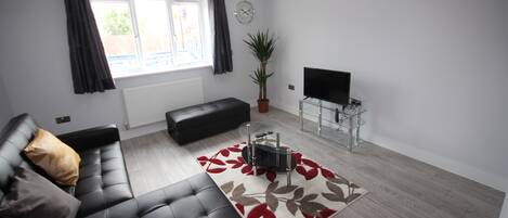 A modern living room with a black leather sofa, a glass coffee table, a red and brown patterned rug, a potted plant, and a flat-screen TV on a glass stand.