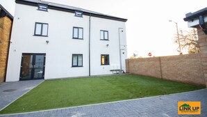 Een modern gebouw met twee verdiepingen, een plat dak, een kleine voortuin met kunstgras en een bakstenen muur aan de rechterkant.