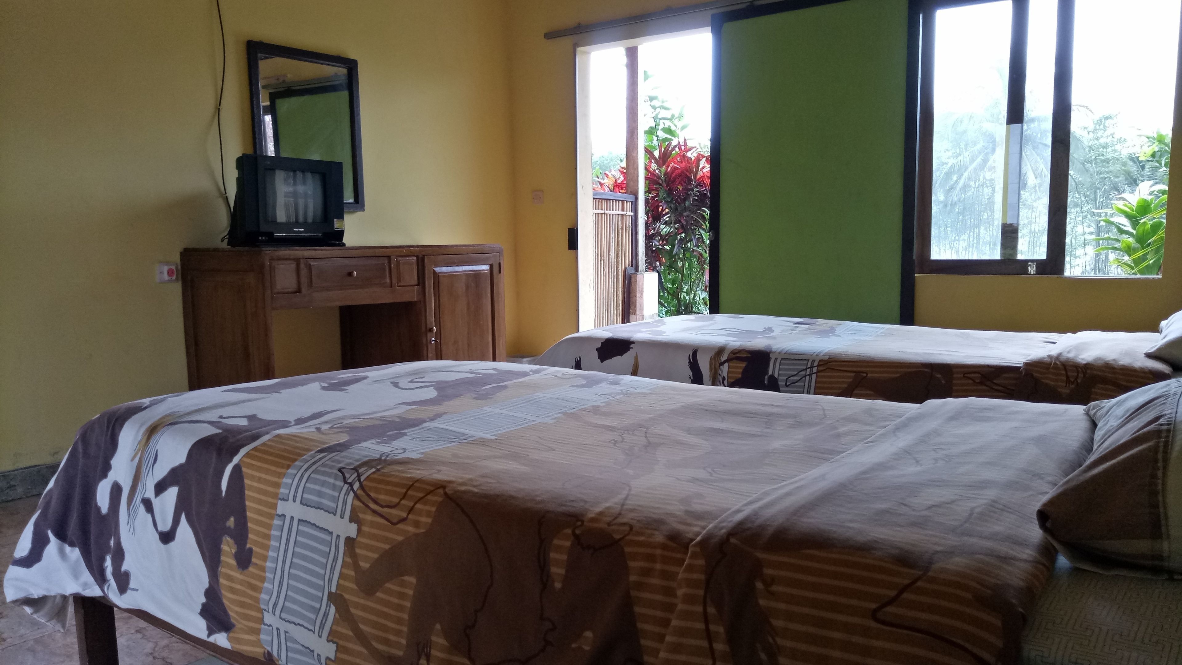 A room with two beds, a wooden desk with a television, and a window with a view of greenery.