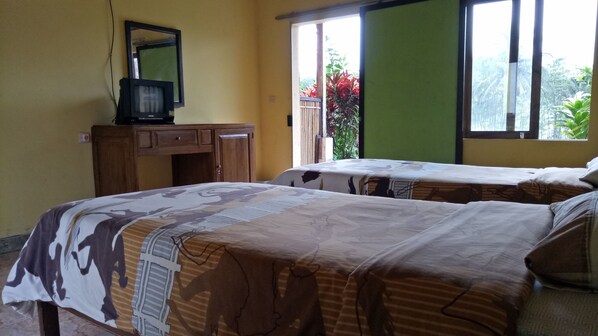A room with two beds, a wooden desk with a television, and a window with a view of greenery. - Asparin Homestay (Banyuwangi)