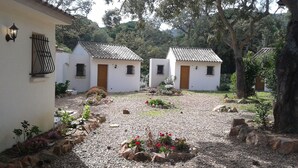 Un patio con tres edificios blancos, cada uno con techo de tejas y puerta de madera. Hay macizos de flores con flores rojas y un sendero de grava.