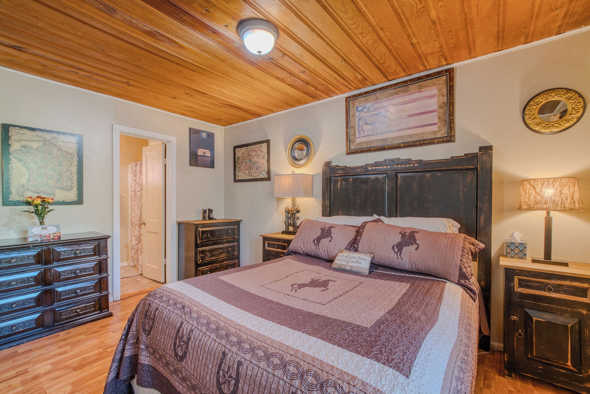 Ein Schlafzimmer mit Holzbalkendecke, einem großen Bett mit gemustertem Überwurf, einem Nachttisch mit Lampe, einem Schminktisch und einem Spiegel an der Wand.