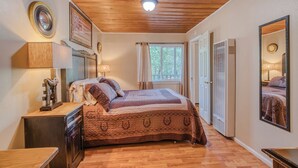 A bedroom with a wooden ceiling, a bed with a patterned bedspread, a nightstand, a mirror, and a window with curtains.