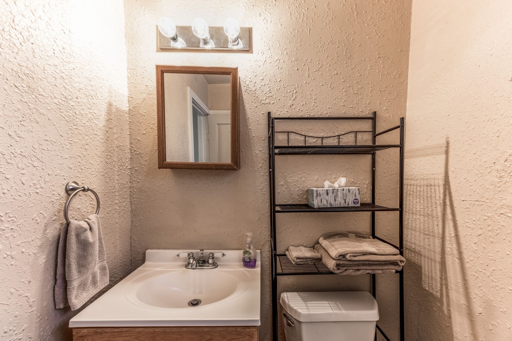 Un bagno con un lavandino bianco, uno specchio sopra, un porta asciugamani, uno scaffale con articoli da bagno e un water.