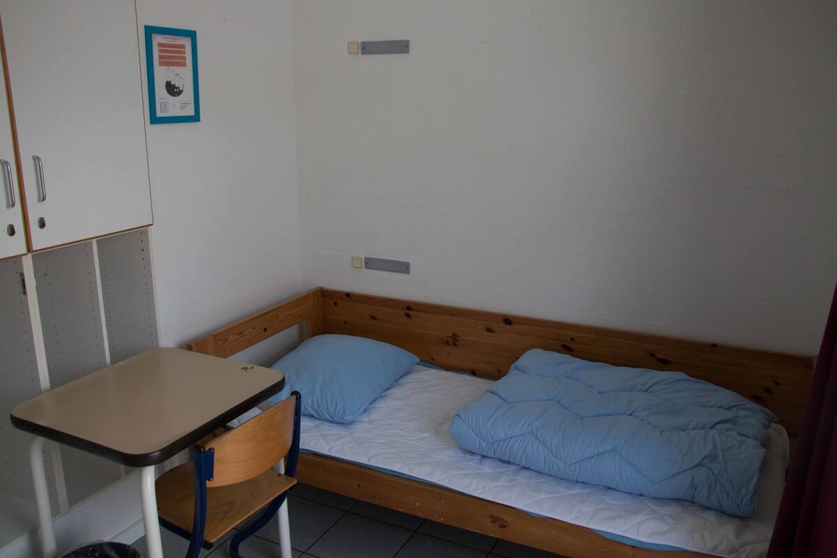 Ein Zimmer mit einem Einzelbett, einem Holzkopfteil, einem kleinen Schreibtisch und einem Stuhl.