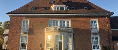 A large brick building with a central entrance, multiple windows, and a small balcony.