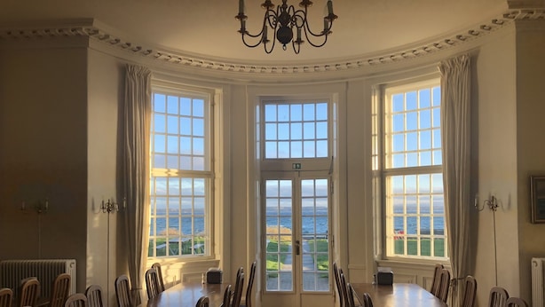 A dining room with a large wooden table and chairs, large windows with a view of the sea, and a chandelier. - Danhostel Helsingør Vandrerhjem (Helsingor)