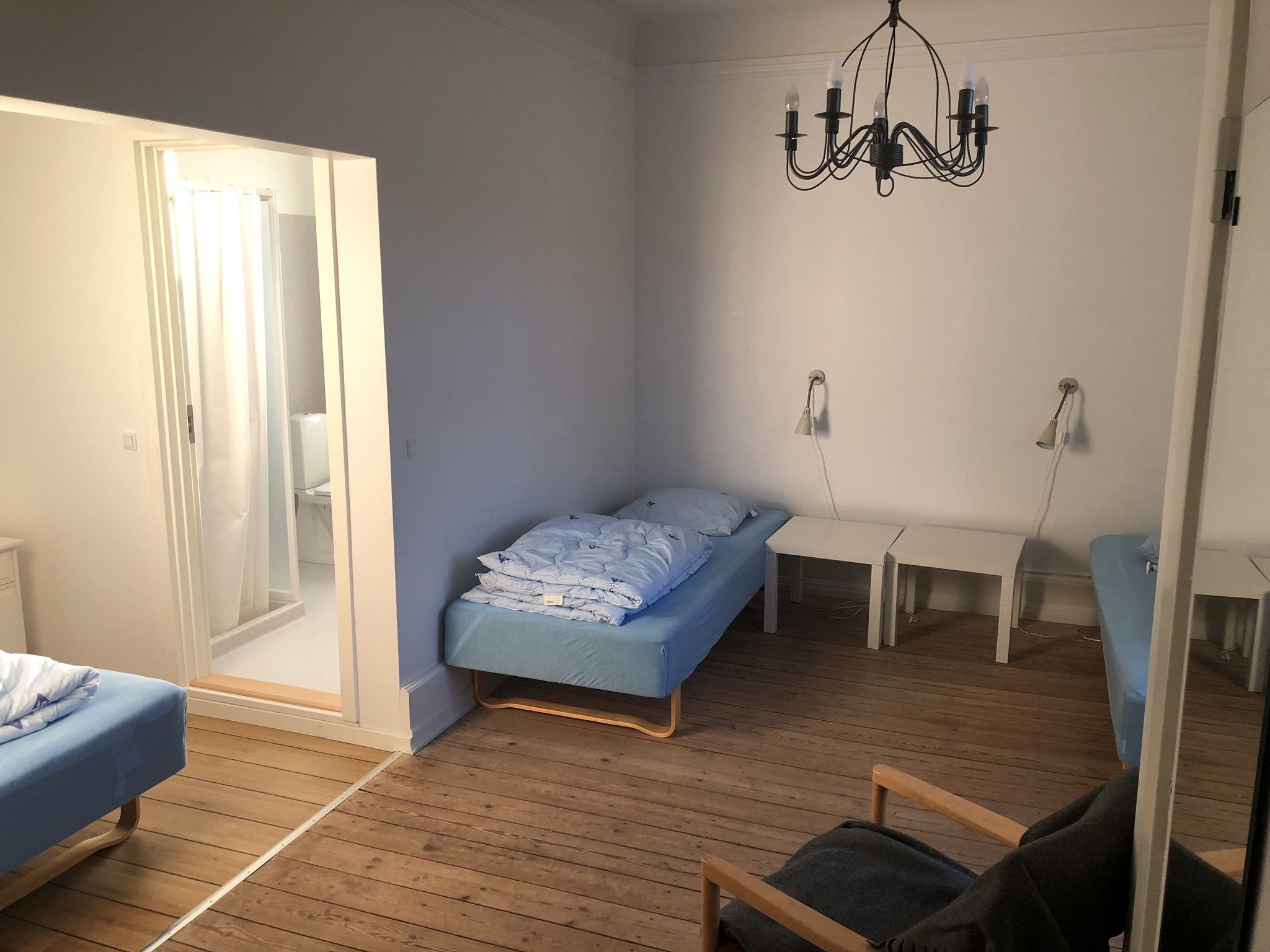 A small, well-lit room with a bed, a chair, a desk, and a chandelier.