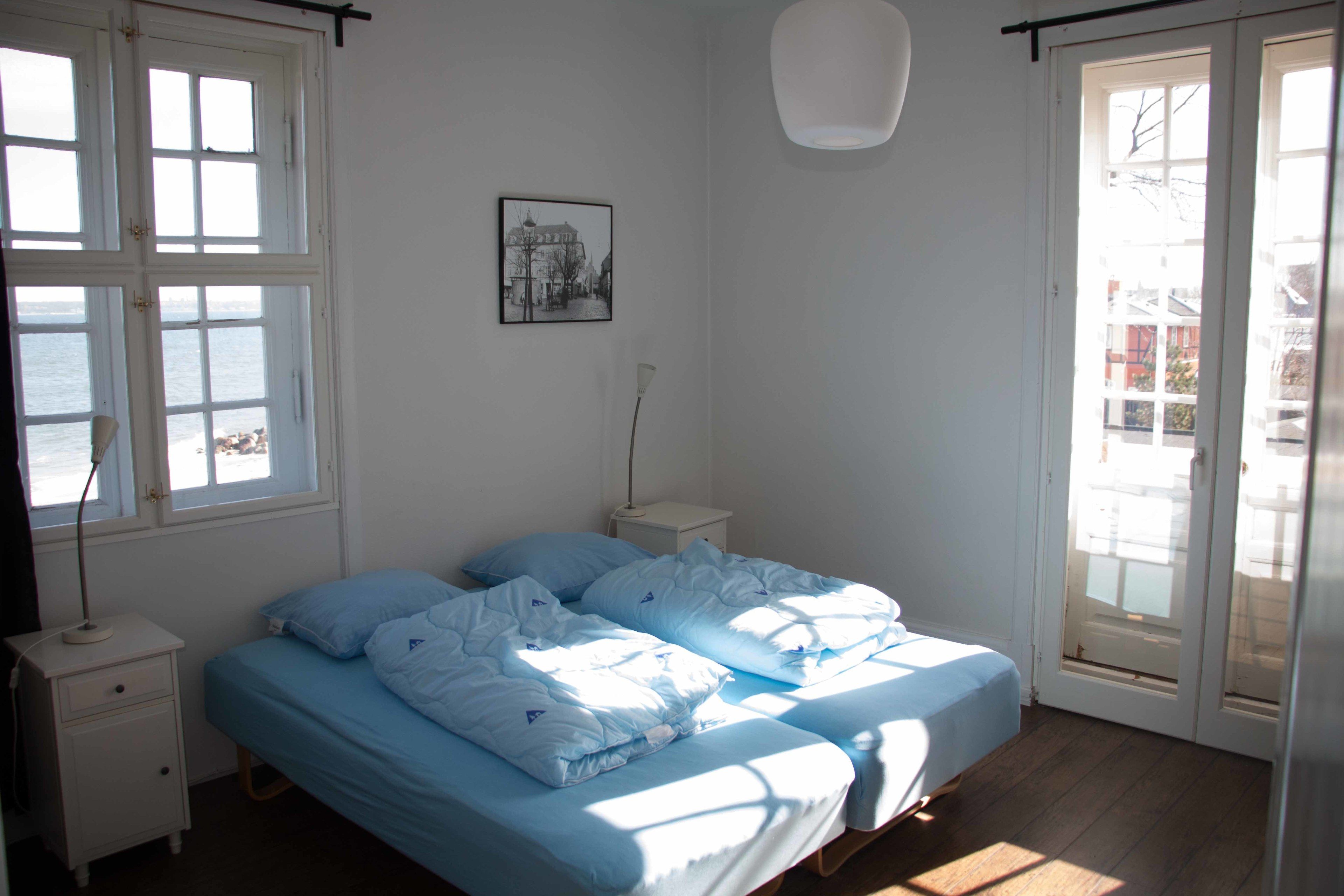 A bedroom with a bed, a nightstand, a lamp, a picture on the wall, and a view of the sea through the window.