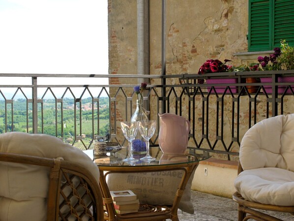 Un balcón con sillas de mimbre, una mesa de vidrio y vistas a un paisaje verde.