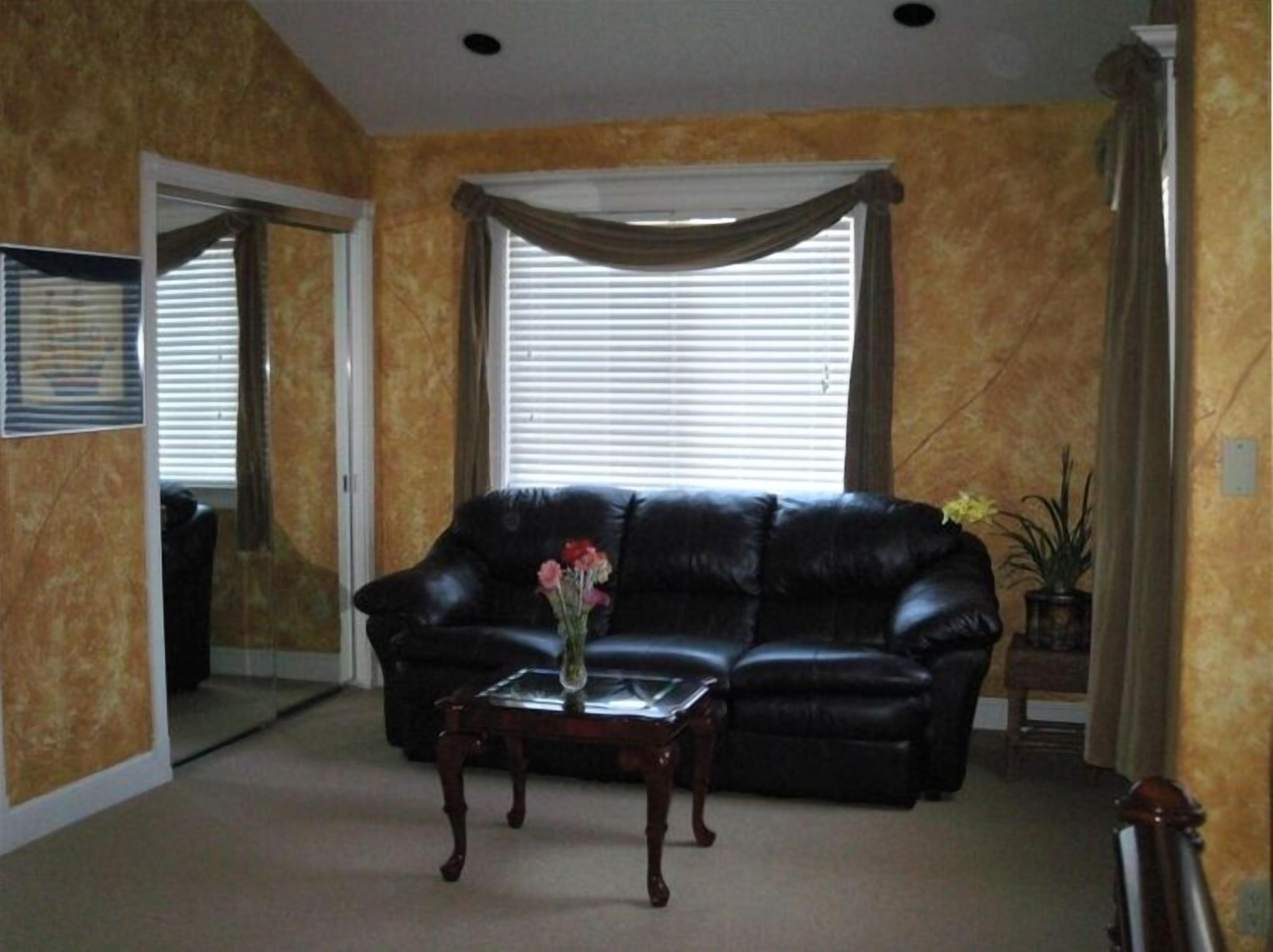 A living room with a black leather sofa, a glass coffee table with a vase of flowers, and a large window with blinds.