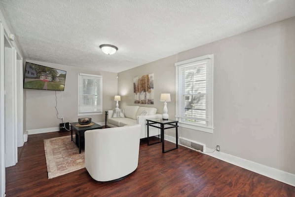 A living room with a sofa, two side tables, a flat-screen TV, and a window with blinds.