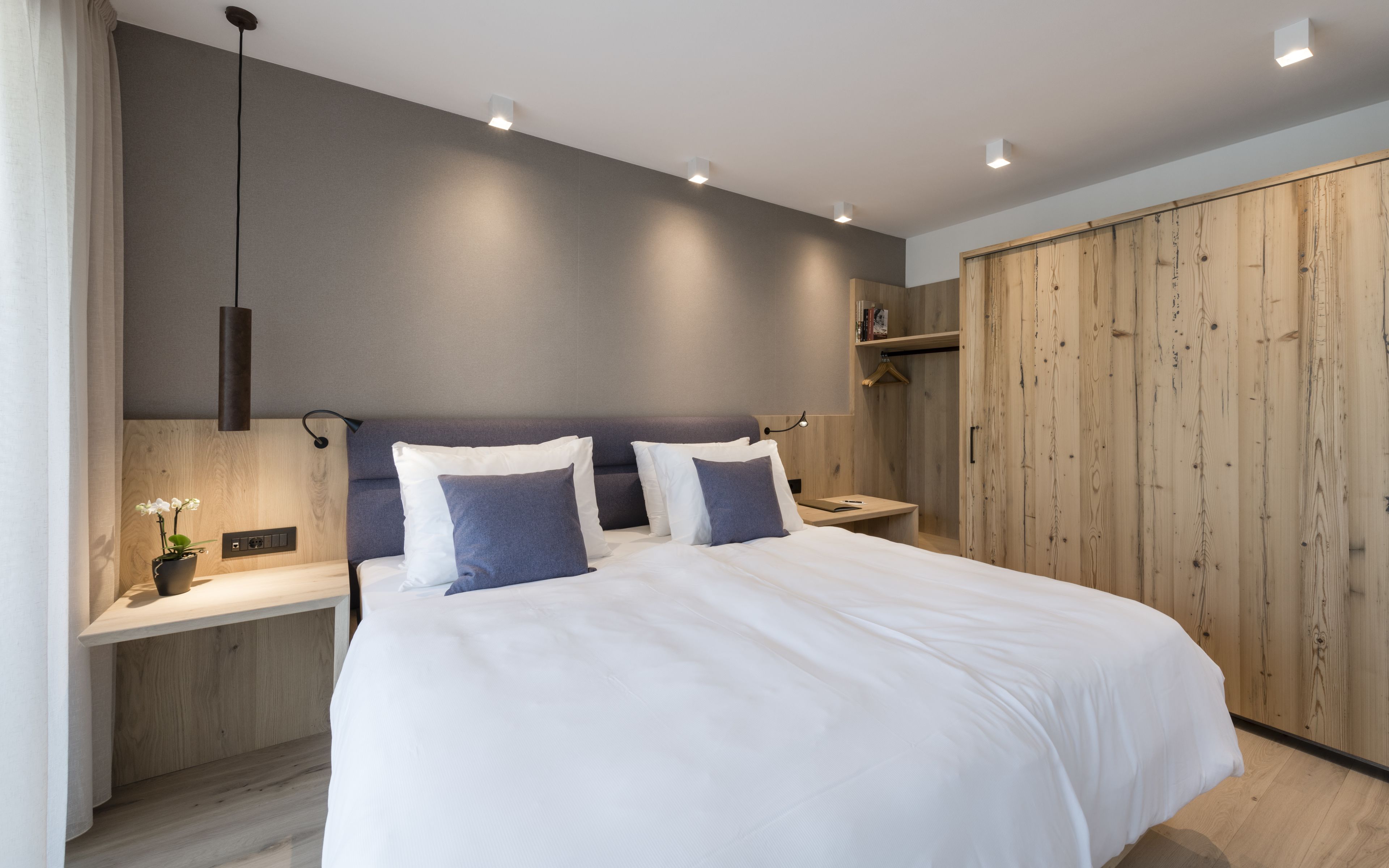 A modern bedroom with a large bed, wooden wardrobe, bedside tables, and a hanging plant.