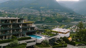 A modern multi-story hotel with a rooftop pool, surrounded by lush greenery and mountains. - Vinea - Suites & Apartments (Tirolo)