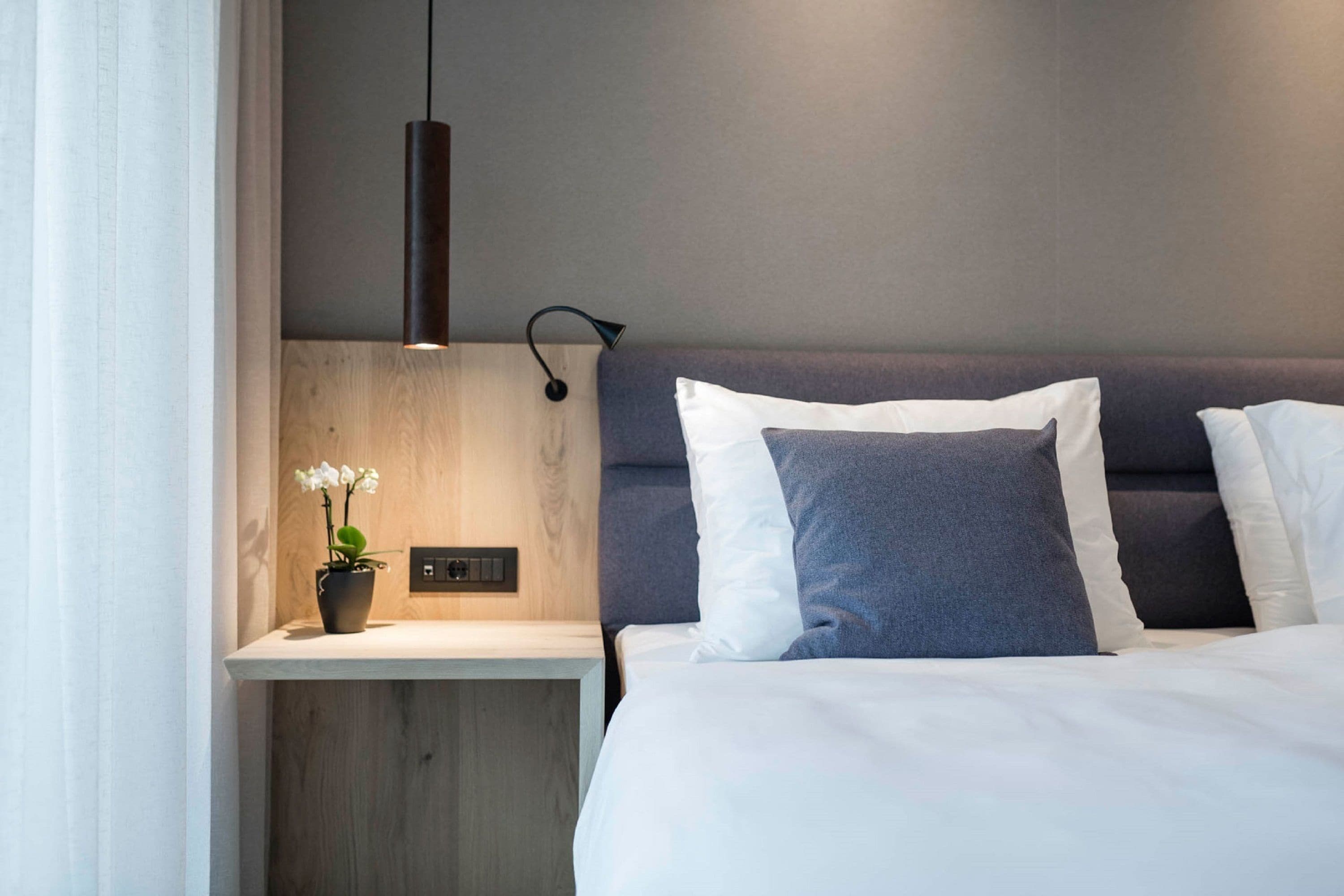 Et moderne hotelværelse med et trænatbord, en potteorkidé, en sengetabletlampe og en seng med hvid og grå sengetøj.