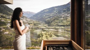 A woman in a white dress standing by a window overlooking a valley with vineyards and mountains. - Vinea - Suites & Apartments (Tirolo)