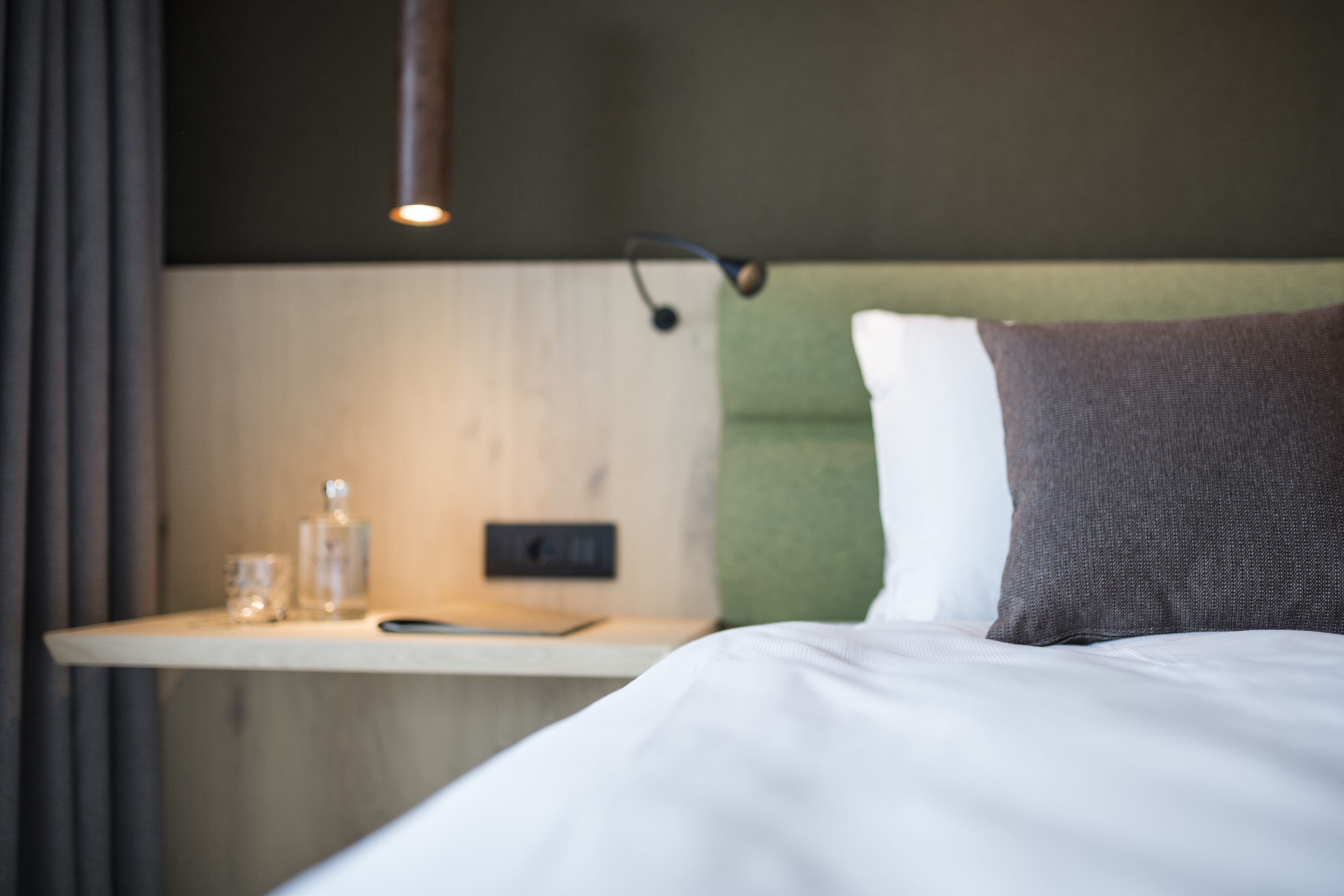 Una cama bien hecha con un almohadón, una mesita de noche con un vaso y una botella, y una lámpara de pared.