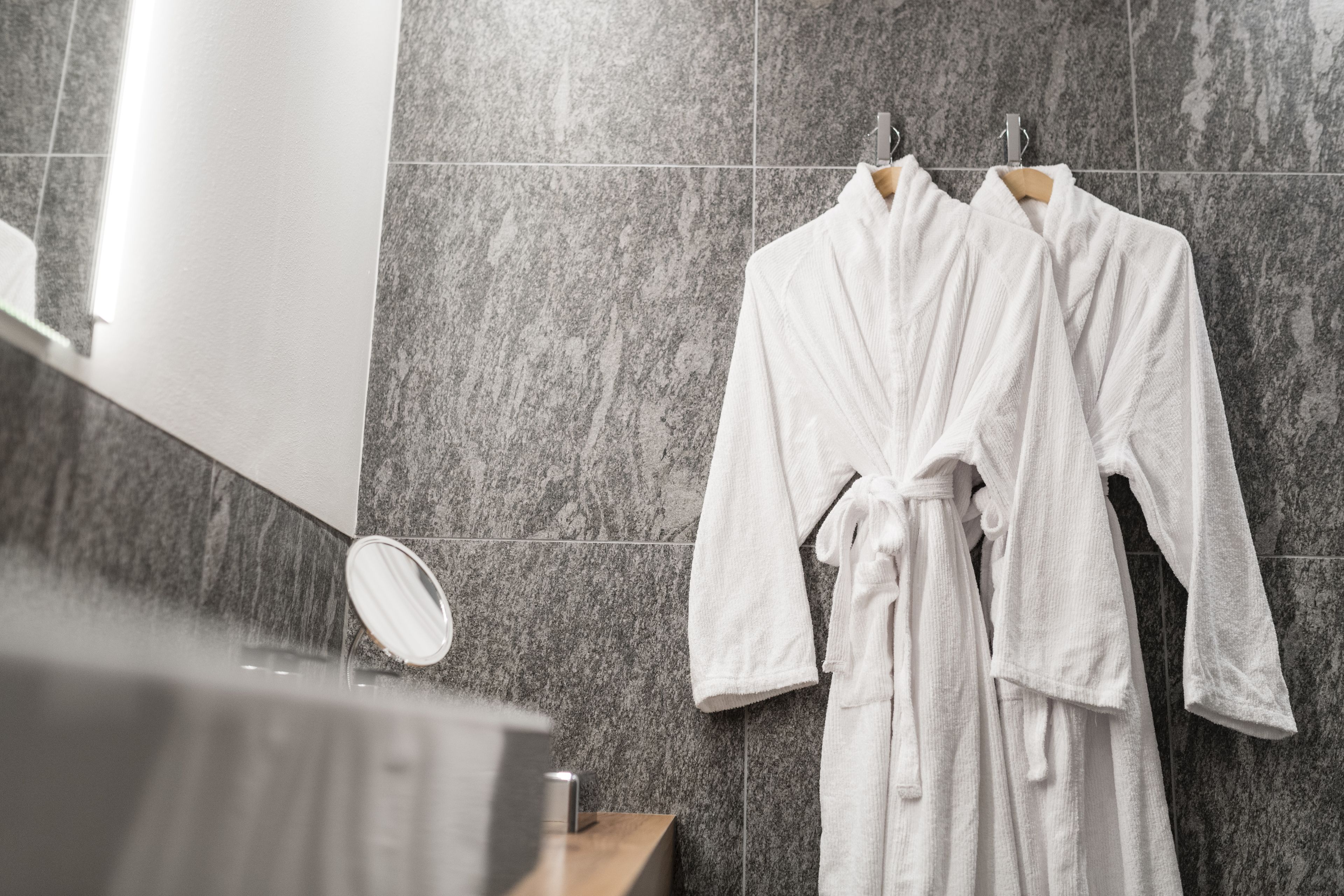 A bathroom with two white bathrobes hanging on a rack, a wooden counter, and a mirror.
