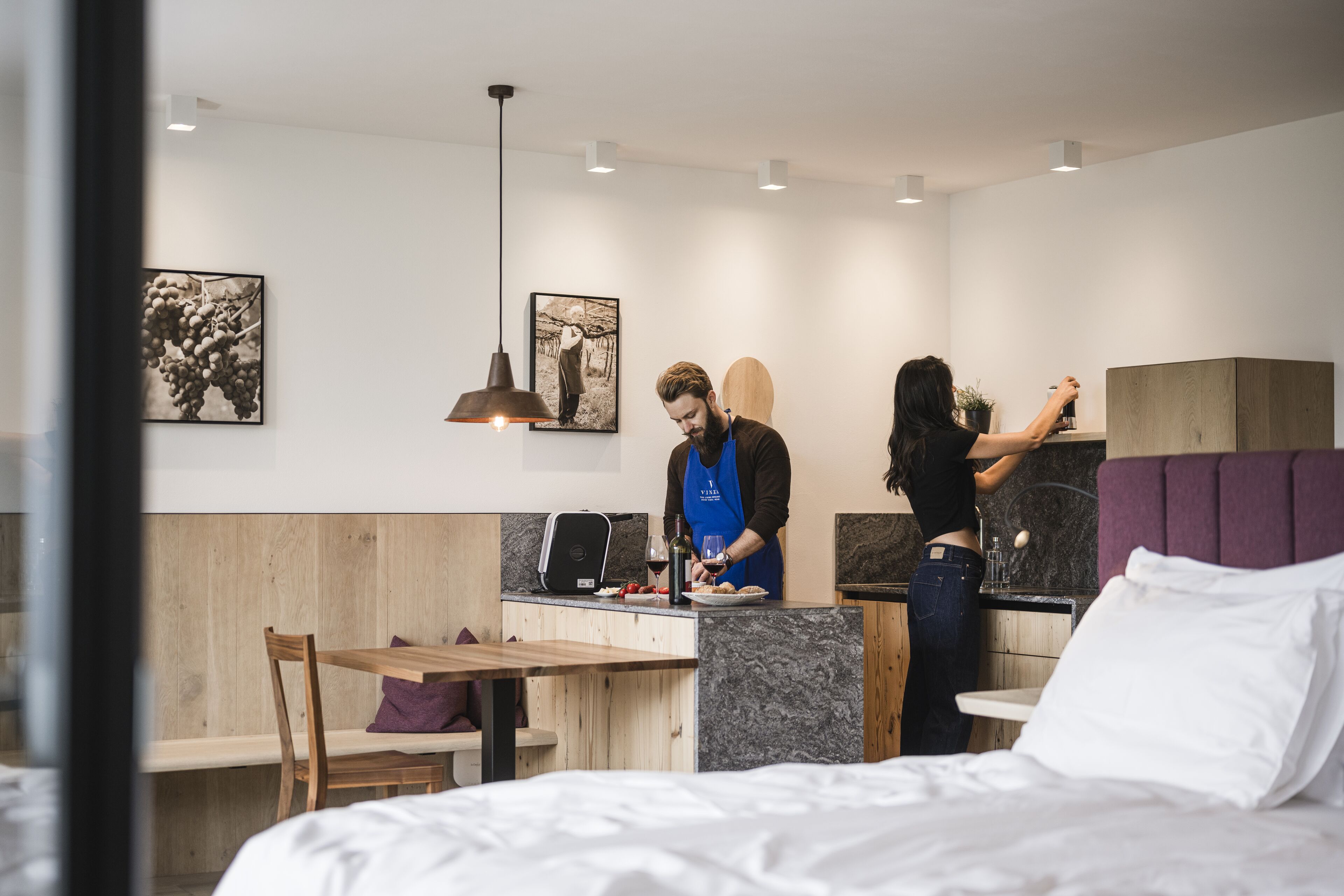 A modern hotel room with a wooden dining table, a bed with white linens, a purple headboard, and artwork on the walls.