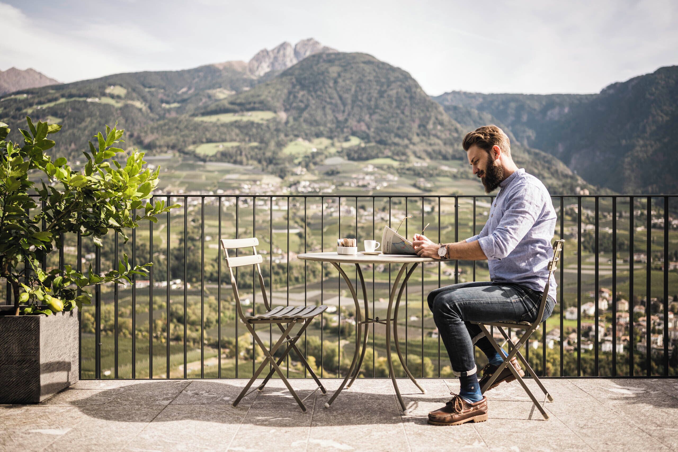 Un hombre sentado en una mesa con una computadora portátil en un balcón con vistas a un paisaje montañoso.