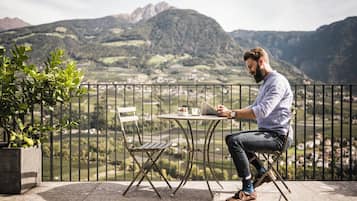 Un hombre sentado en una mesa con una computadora portátil en un balcón con vistas a un paisaje montañoso.