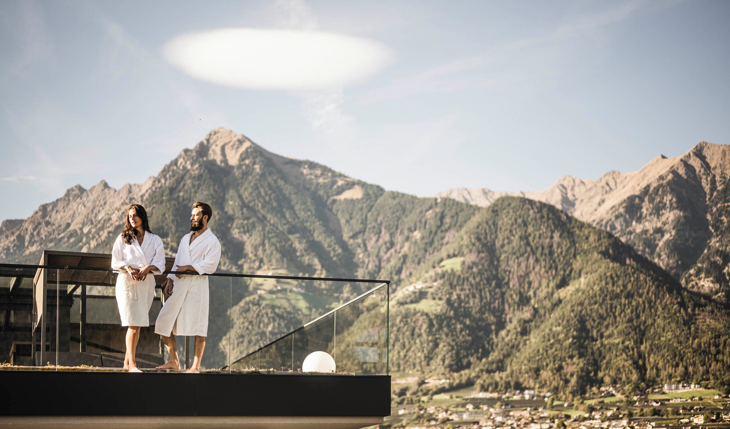 Two people in white robes standing on a balcony with a mountain backdrop.