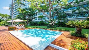 A swimming pool with lounge chairs and a wooden deck, surrounded by lush greenery and a multi-story building. - Avalon - 907 (Puerto Vallarta)