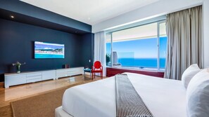A modern hotel room with a large bed, a flat-screen TV mounted on the wall, a red chair, and a large window offering a view of the ocean.