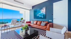 A modern living room with a sofa, coffee table, and chairs. A large painting of a wave is on the wall.