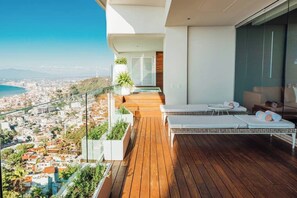 Un balcon doté d’un plancher en bois, de fauteuils de jardin blancs et d’une balustrade en verre offrant une vue sur le paysage urbain.