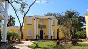 A yellow building with a black door and white columns, surrounded by a well-maintained lawn and trees. - C5, Flagler location in downtown, close to beaches, private patio, free parking! (St. Augustine)