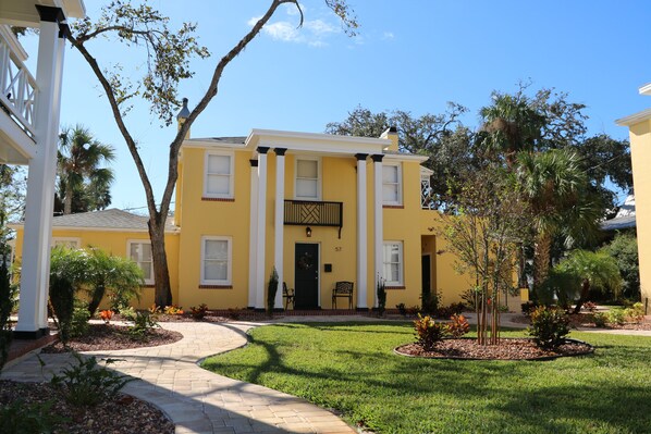 A yellow building with a black door and white columns, surrounded by a well-maintained lawn and trees.