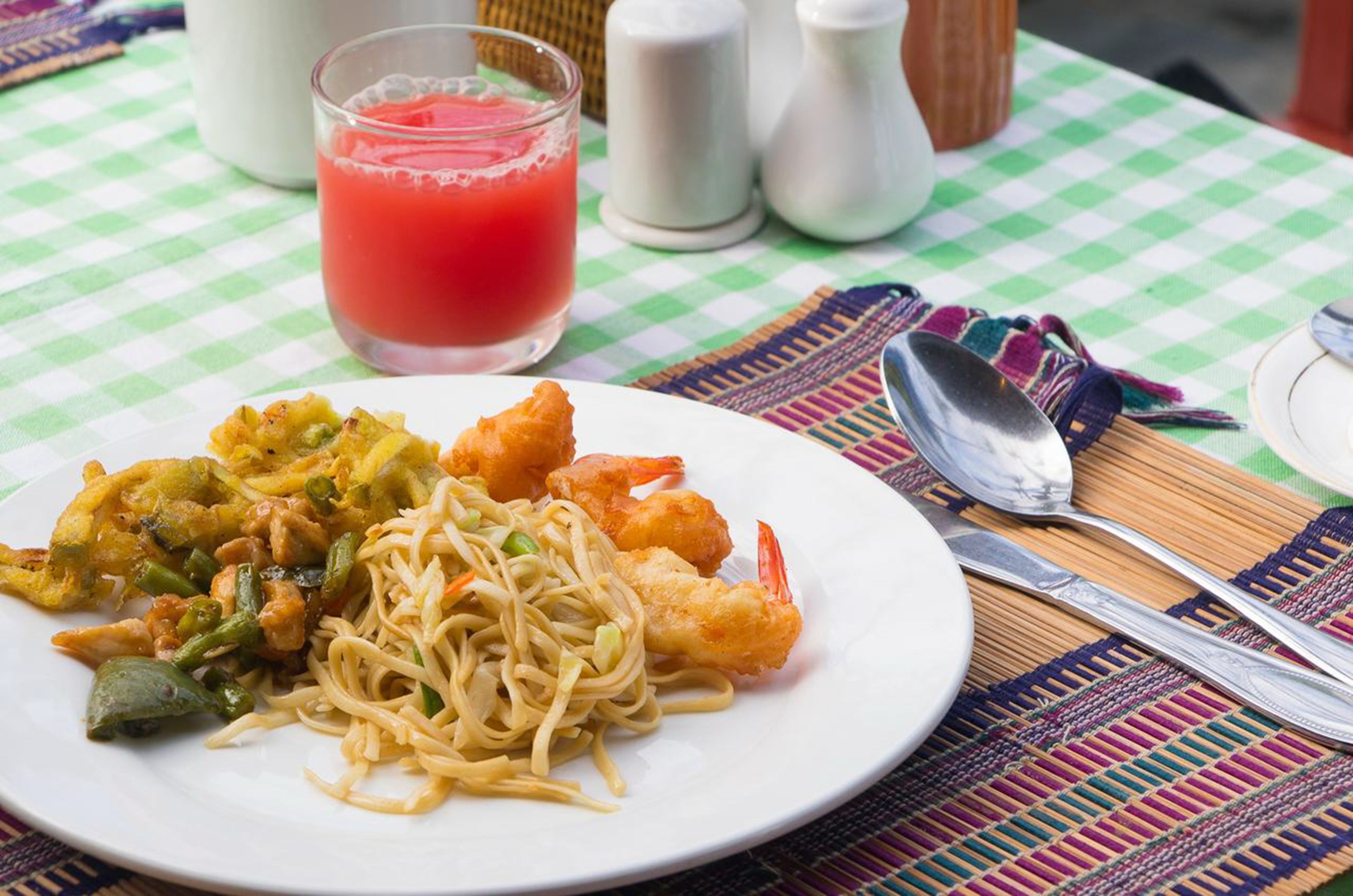 Prato de comida com macarrão e alimentos fritos, um copo de bebida vermelha e talheres sobre toalha de mesa xadrez.