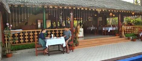 An outdoor dining area with a thatched roof, a pool, and a wooden fence.
