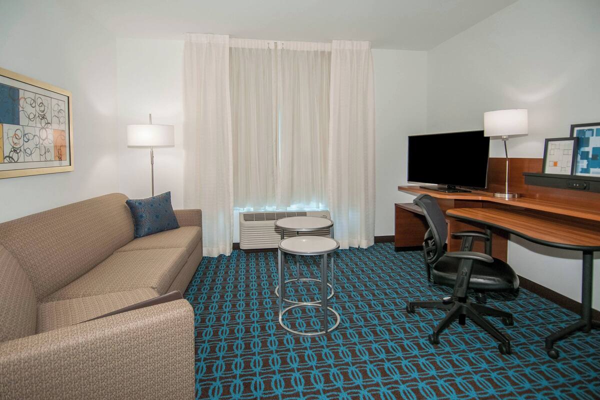 A hotel room with a brown sofa, a desk with a computer and a TV, a small round table, and a patterned carpet.