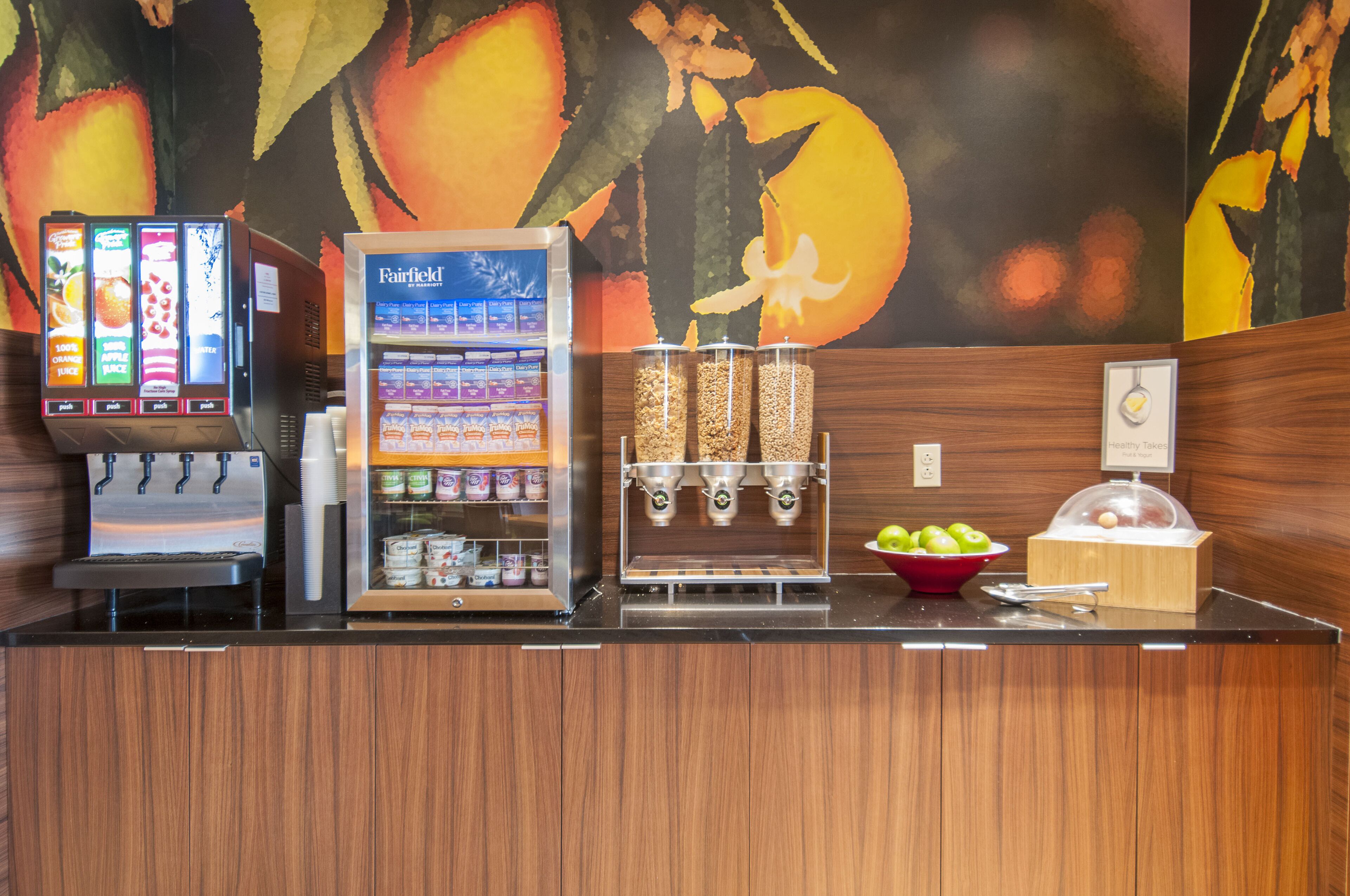A counter with a juice dispenser, a refrigerated display case, and a bowl of apples.