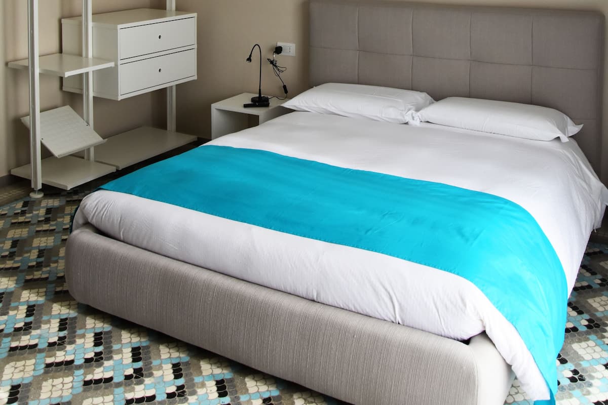 A hotel room with a bed, a white bedside table, a lamp, a wardrobe, and a patterned floor.