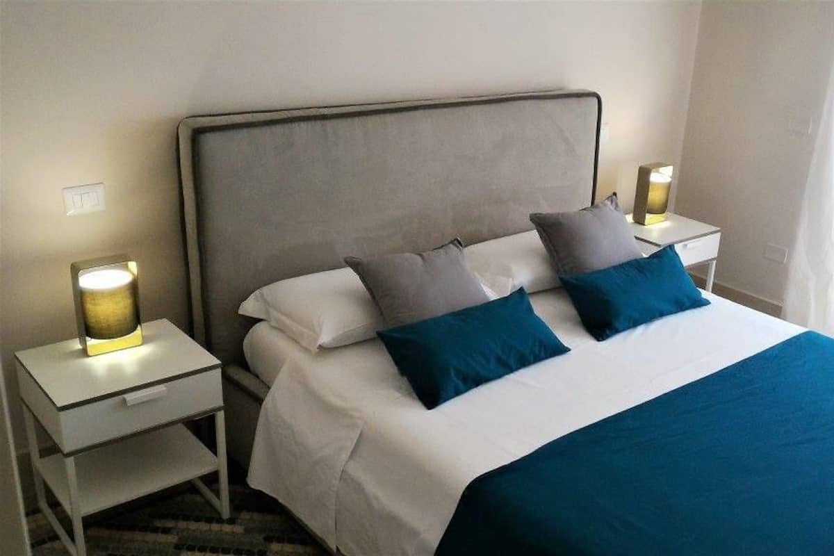 A neatly made bed with a grey headboard, white bedding, and two blue pillows. There are bedside tables with lamps on either side of the bed.