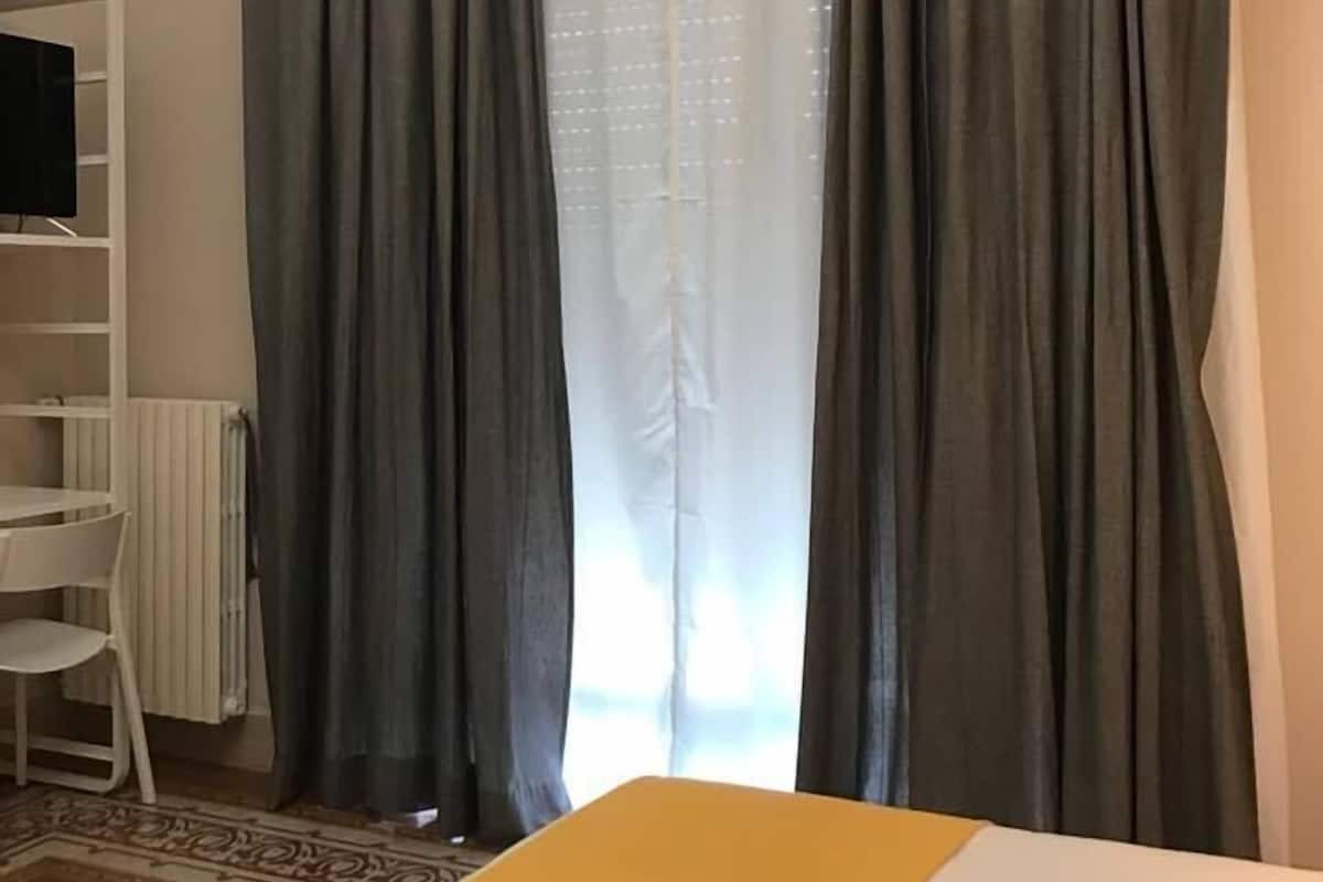 A hotel room with a bed, a white radiator, a white chair, and a window with grey curtains.