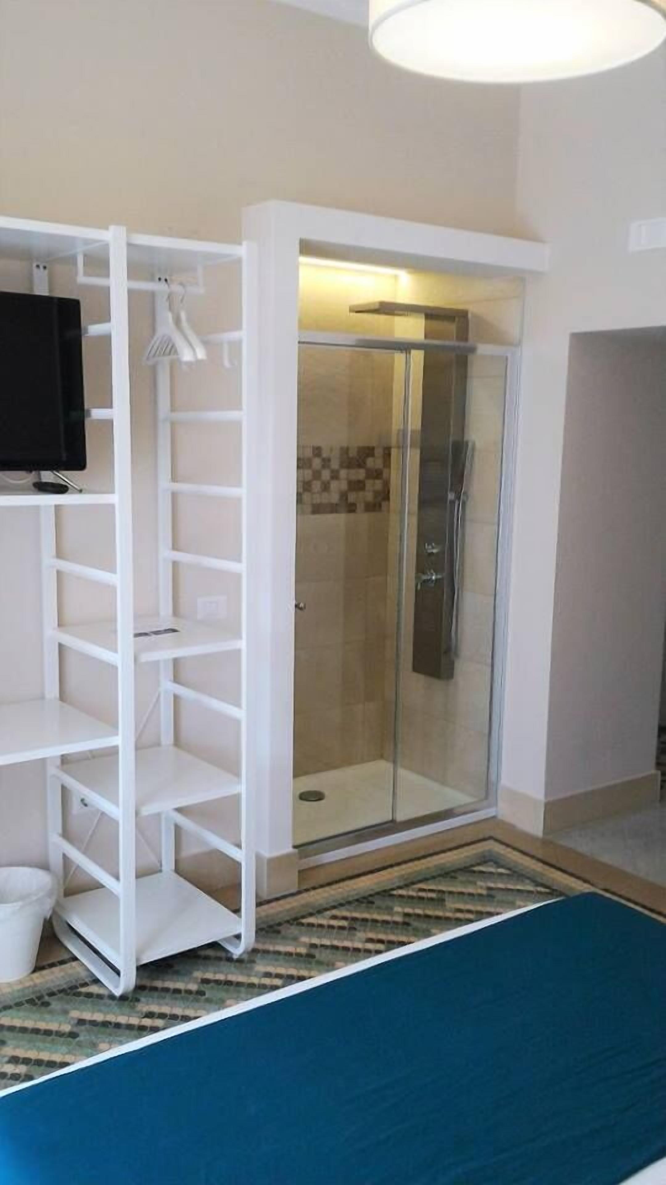 A hotel room with a walk-in shower, a white shelving unit, and a flat-screen TV.