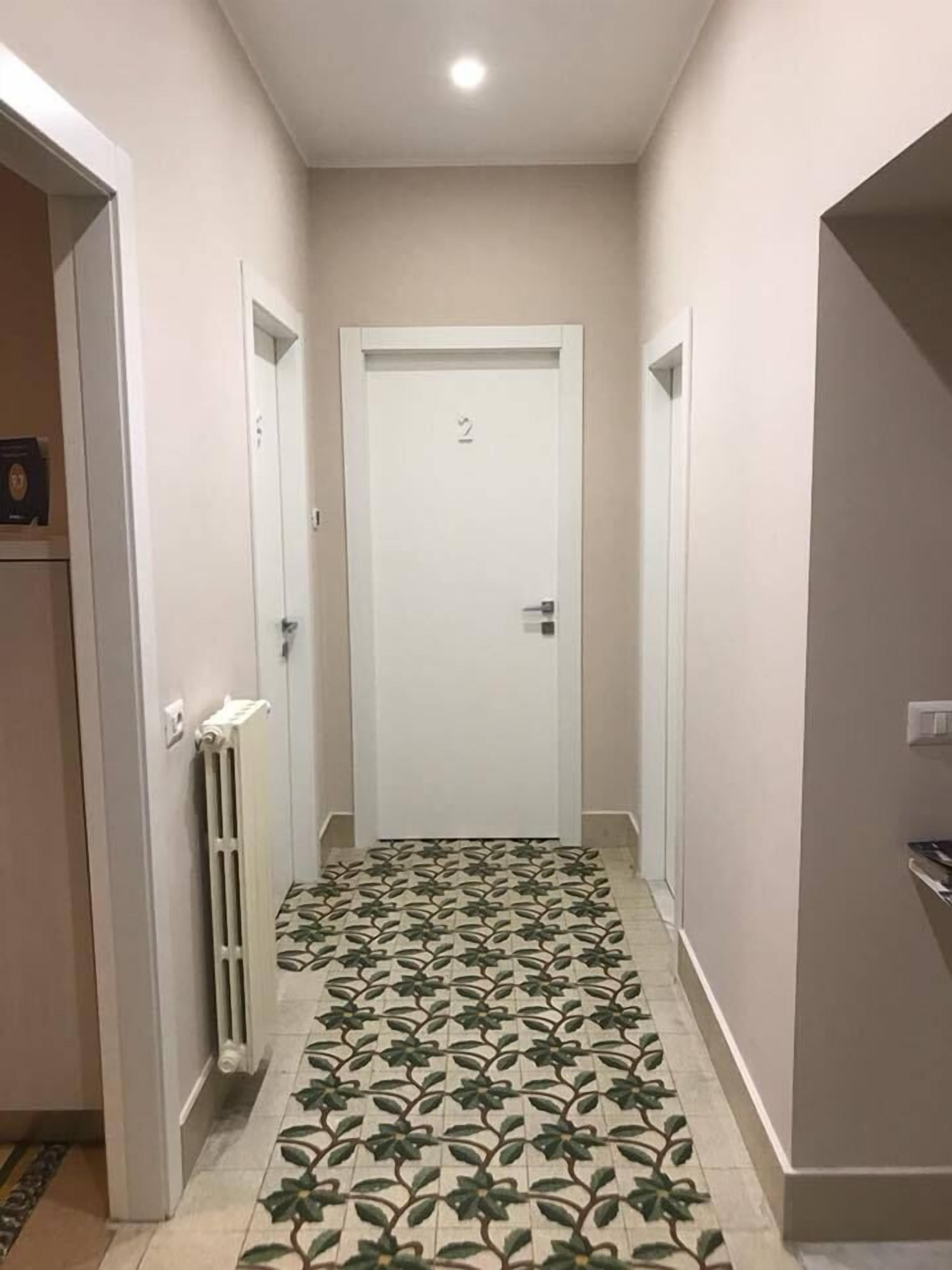 Un pasillo estrecho con piso estampado, una puerta blanca con un número y un radiador a la izquierda.