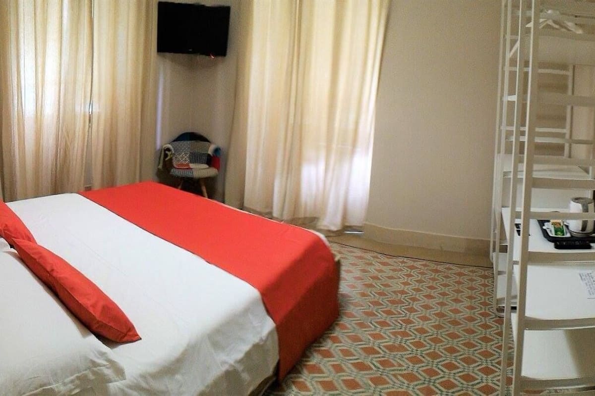 A hotel room with a bed, a TV mounted on the wall, a small table with a chair, and a white shelf unit.