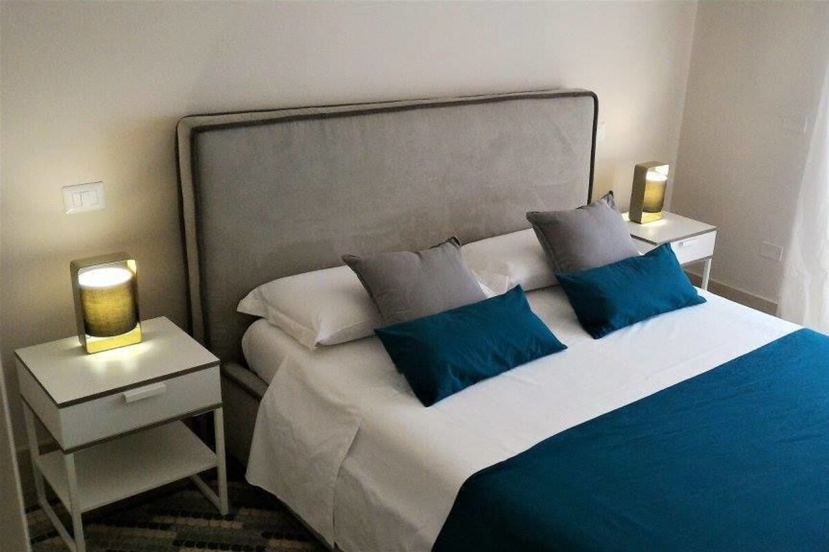 Un lit bien fait, avec une tête de lit grise, deux tables de chevet avec des lampes et une fenêtre avec des rideaux.