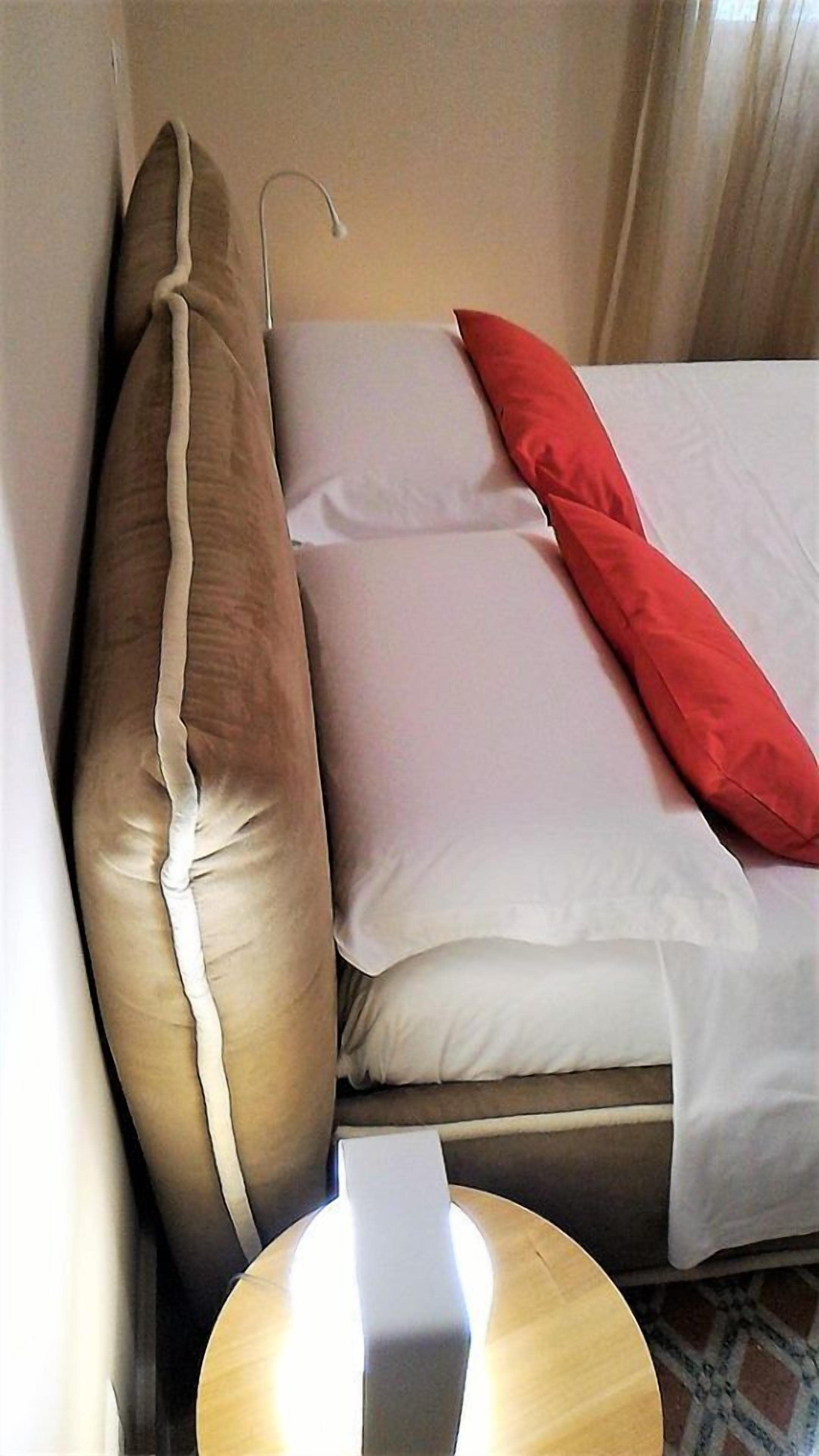 A bed with white and red pillows, a wooden nightstand, and a hanging lamp.