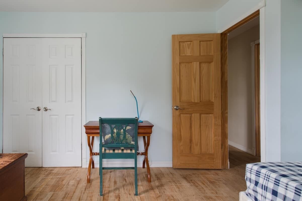A room with a wooden door, a green chair, a small wooden table, and a bed with a checkered bedspread.