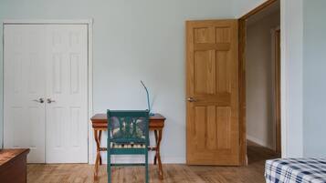 A room with a wooden door, a green chair, a small wooden table, and a bed with a checkered bedspread.