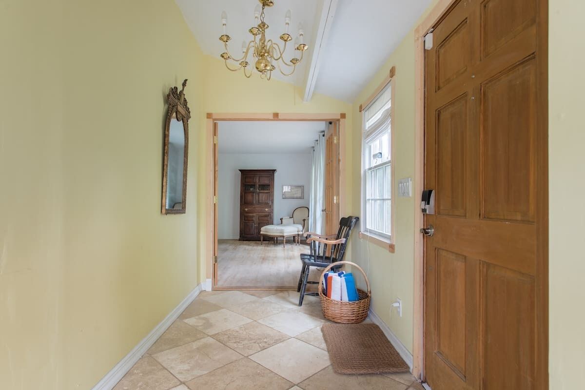 A hallway with a wooden door, a chandelier, a mirror, a chair, and a basket on the floor.