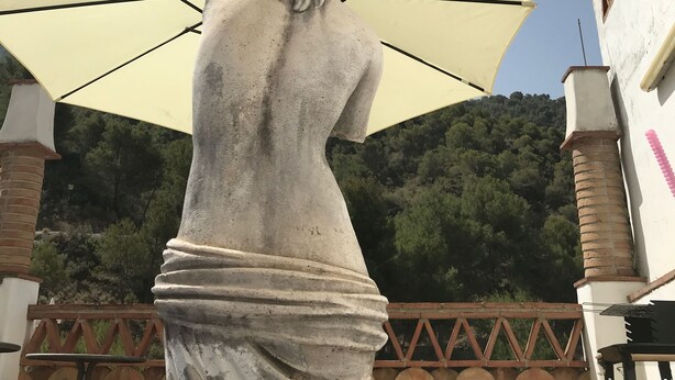 Une statue d’une femme drapée, placée sous un grand parasol, avec une chaîne de montagnes en arrière-plan et un bâtiment doté d’une cheminée en briques.